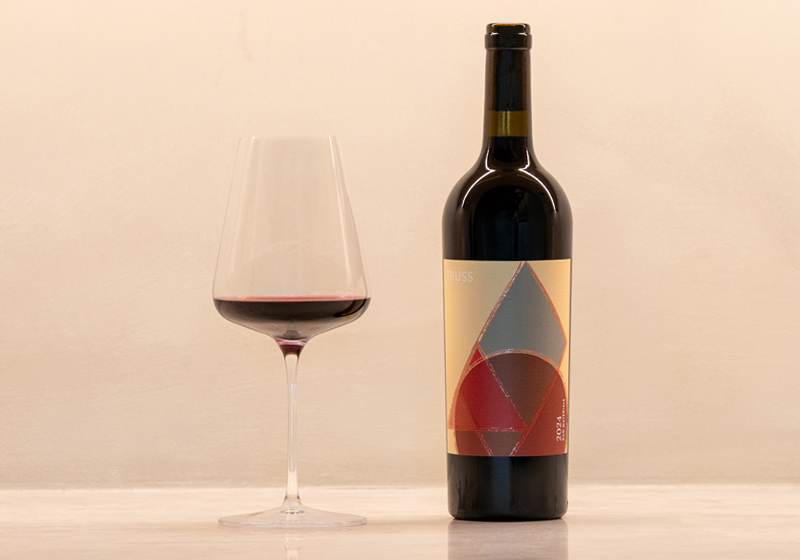 Truss red wine bottle with glass of red wine in nice tasting room setting.