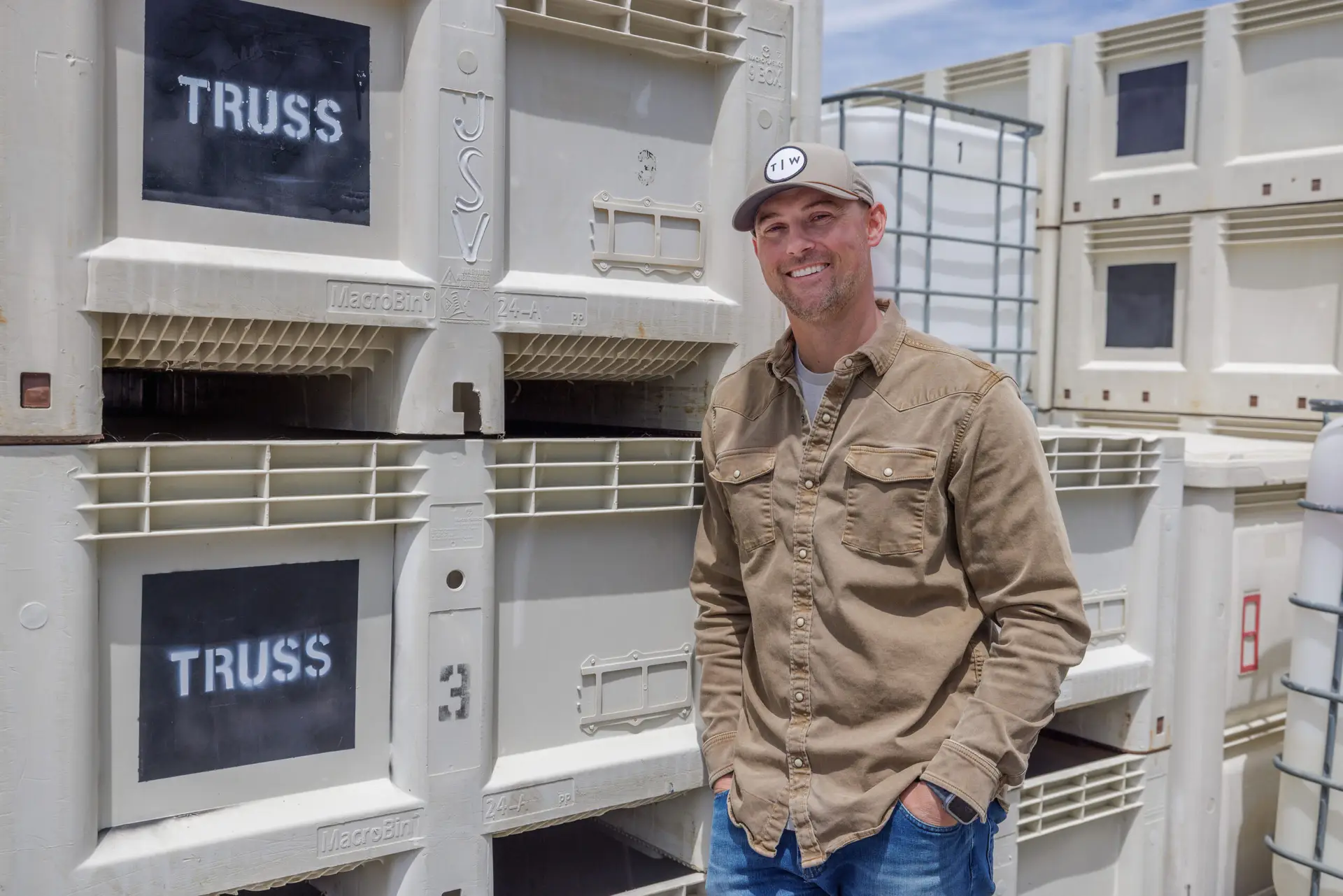 Man in a tan shirt and cap smiling while standing beside large white harvest bins labeled with the Truss logo outdoors at a winery.